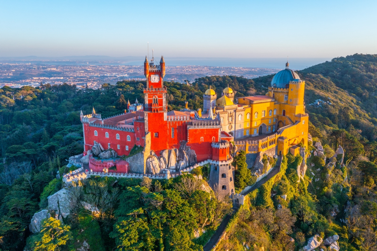 Aerial view of Pena Palace's red and yellow towers on a wooded, rocky hilltop, with the plain stretching away to the coast