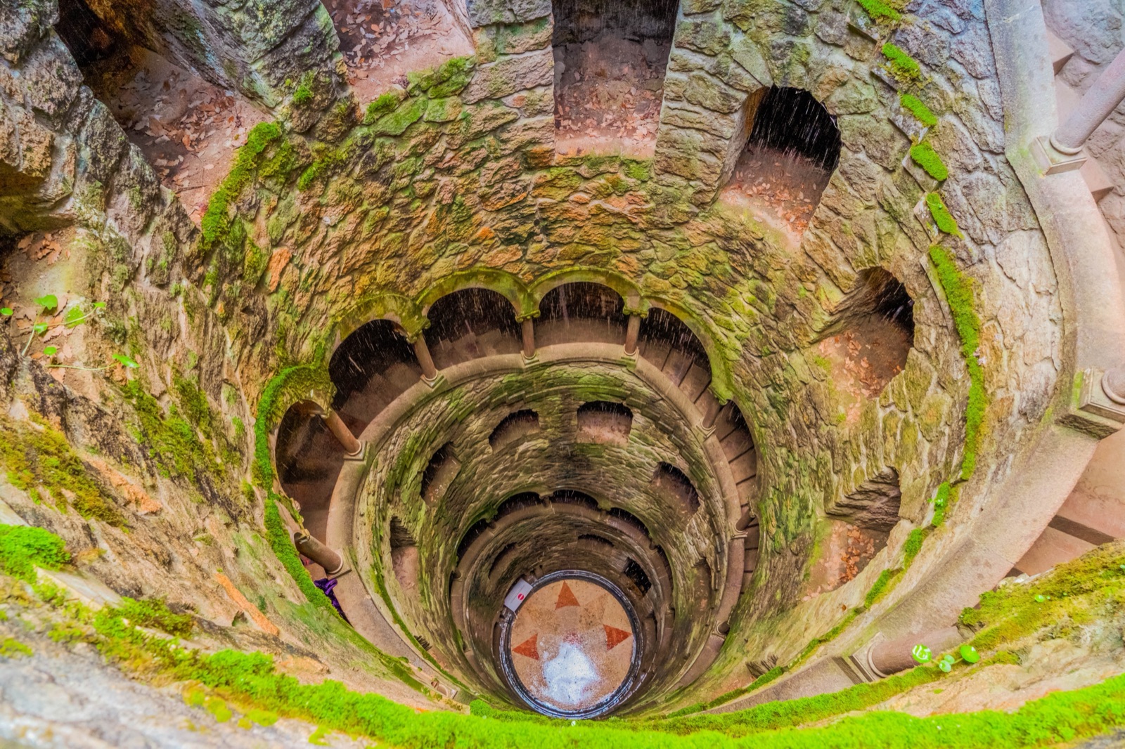 Looking down the mossy spiral staircase of the Initiation Well at Quinta da Regaleira to a patterned floor far below